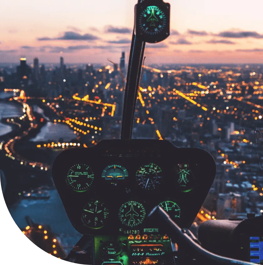 View from a helicopter cockpit at dusk over a lit city skyline, with the instrument panel in the foreground